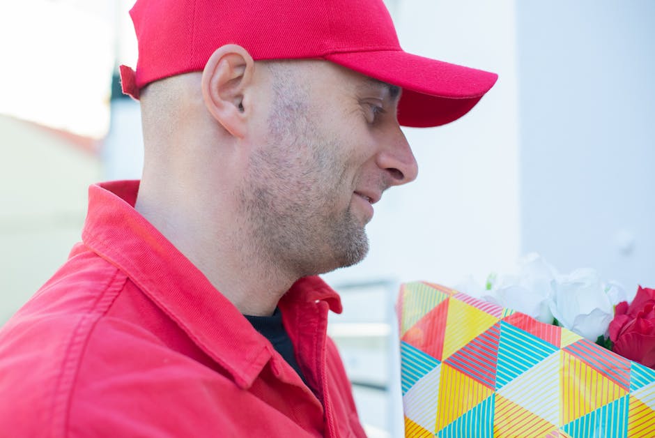 A man wearing a red cap and matching red jacket is holding a bouquet of fresh flowers wrapped in colorful, geometric-patterned paper, with visible white and pink blossoms. He is outdoors, and the back