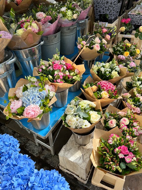 A display of fresh flower bouquets arranged in metal buckets and wrapped in brown paper, featuring a variety of pastel-colored flowers including pink roses, white hydrangeas, blue hydrangeas, and carn