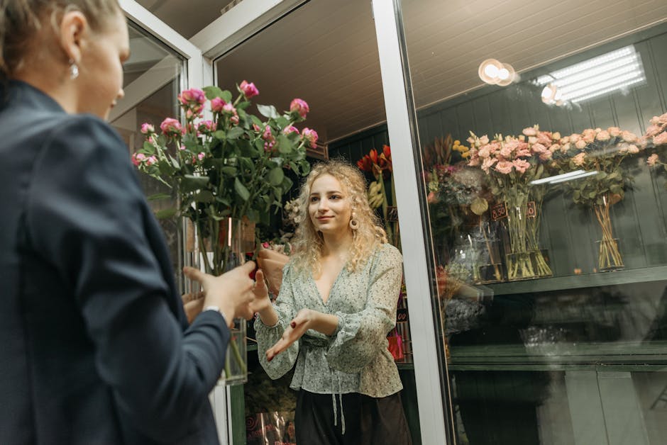 A female customer with curly blonde hair, dressed in a light patterned blouse, is selecting a bouquet of fresh pink roses wrapped in clear plastic from a florist shop window. The florist, whose face i