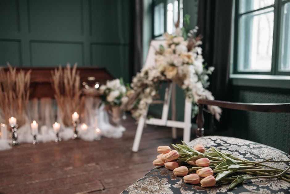 A floral arrangement with soft pink tulips lying on a patterned cushion, featuring smooth petals and green stems, positioned in the foreground of a dimly lit interior space. In the background, a large