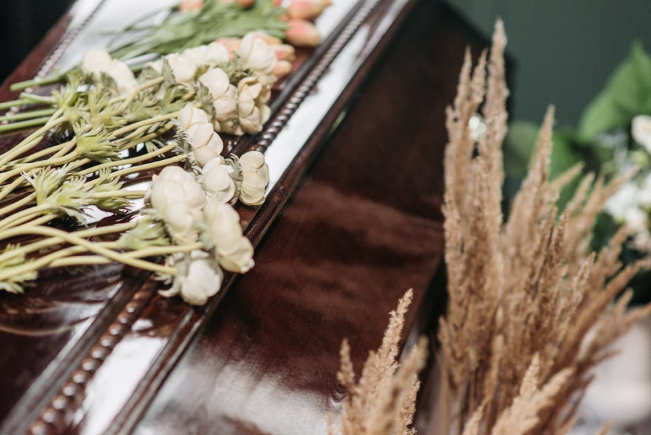 A close-up of a funeral flower arrangement featuring a row of white lisianthus blossoms with delicate, layered petals and slender green stems, laid horizontally on a polished wooden surface. To the ri