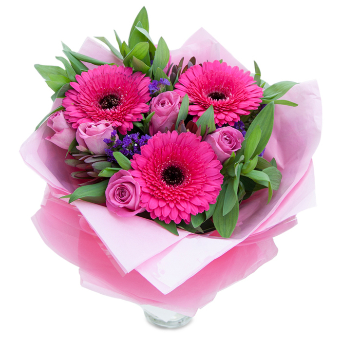 A graceful arrangement that balances bold colour with soft romance, photographed on a white field that lets each bloom read clearly. Three large gerbera daisies in a vivid fuchsia tone form the heart of the bouquet; their concentric petals fan outward around distinct dark centres, offering a striking graphic contrast. Scattered among them are several lavender-pink roses, their slightly muted, dusty-rose petals tightly furled in the manner of newly cut blooms, giving an intimate, tender feeling. Flecks of purple statice nestle into the spaces between flowers, their fine, papery clusters bringing airiness and a complementary coolness to the warm palette. Generous green foliage - likely salal with touches of eucalyptus - frames the composition, broad leaves providing a lush canvas that amplifies the pinks and supplies a crisp, slightly dewy visual freshness. The bouquet is wrapped with care: an inner layer of soft pale-pink tissue creates a gentle base, while an outer sheet of translucent fuchsia-pink cellophane with a subtle crinkle produces a delicate shimmer and a finished, gift-ready look. A clear glass vase rim shows at the bottom, indicating the bouquet is arranged for immediate display on a mantle or centrepiece. The scent profile is understated and pleasing - roses offering a classic floral perfume and gerberas contributing a faint, green note - and the arrangement conveys warmth, appreciation, and celebration. As a florist in the Bromley Common area, I imagine this piece finding a welcome home at a family birthday, a Mother's Day breakfast, or a small anniversary dinner in nearby Keston or Hayes Common.